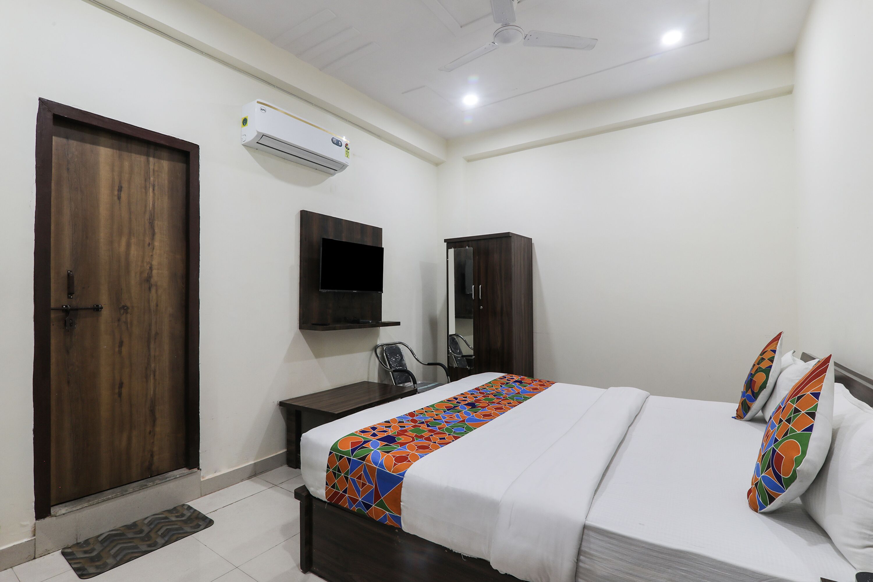 A hotel room with a wooden door, a flat-screen TV mounted on the wall, a desk, and a bed with a colorful bedspread.