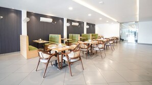 A modern restaurant with wooden tables and green cushioned booths, white ceiling lights, and a tiled floor.