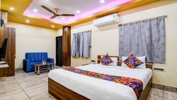 A modern hotel room with a bed, a blue sofa, a small table with drinks, and a ceiling fan.
