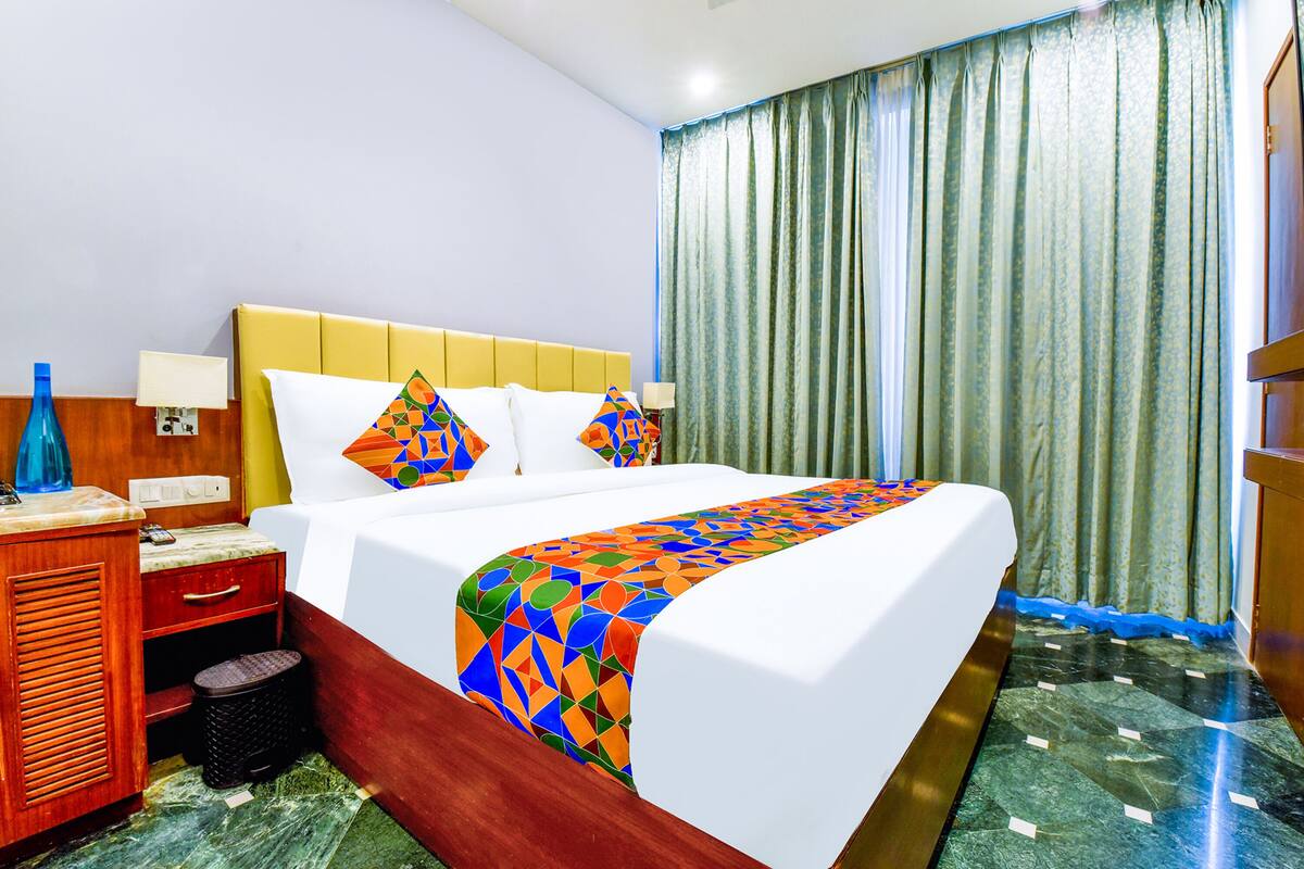 A hotel room with a bed, a wooden nightstand, a lamp, a trash bin, and a window with curtains.