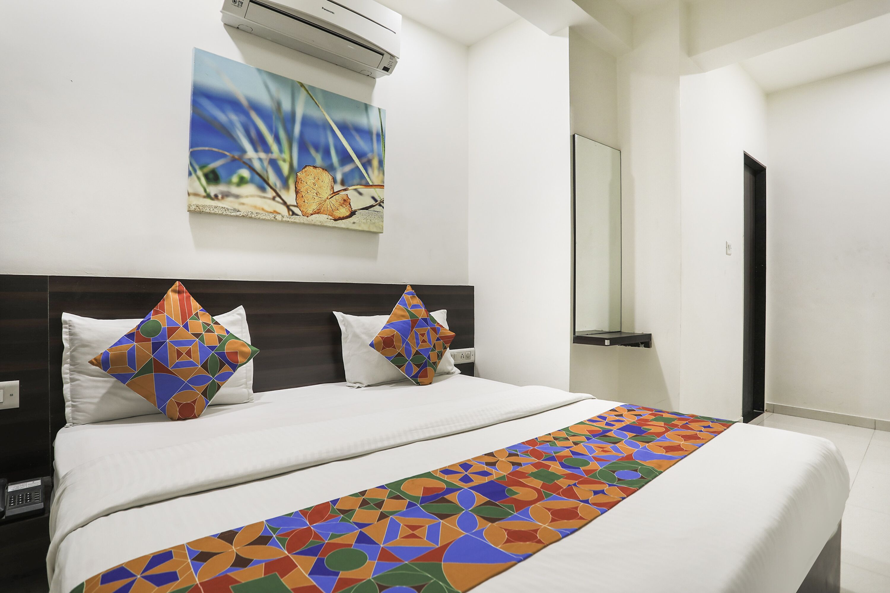 A hotel room with a large bed, two pillows, a colorful bedspread, a painting on the wall, and a mirror.