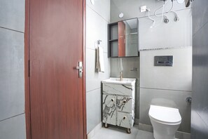 A modern bathroom with a wooden door, a marble vanity, a white toilet, and a mirror cabinet. - FabHotel Embrace (New Delhi)