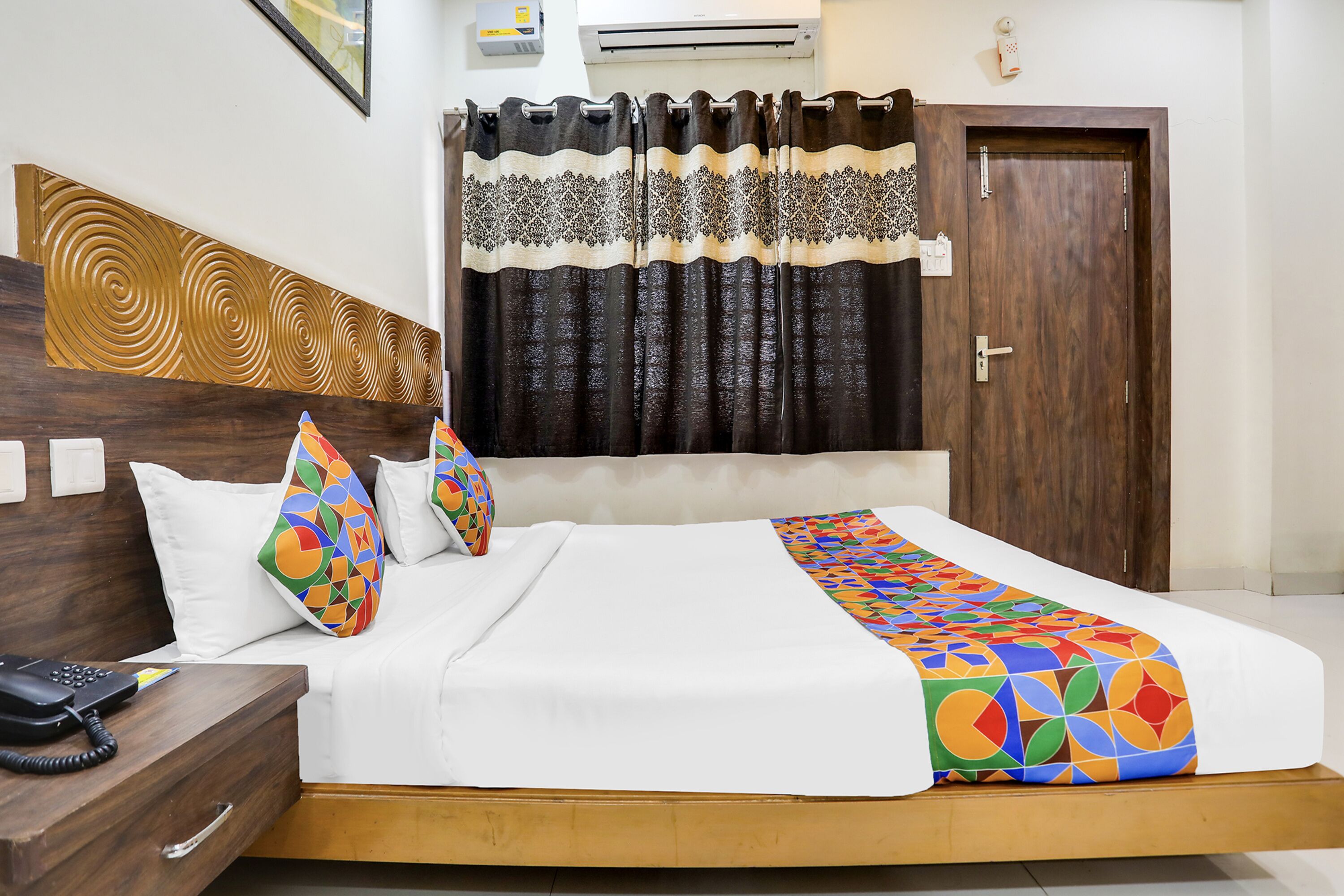 A hotel room with a wooden headboard, a bed with white bedding and colorful geometric pillows, a bedside telephone, and a curtain with a black and white pattern.