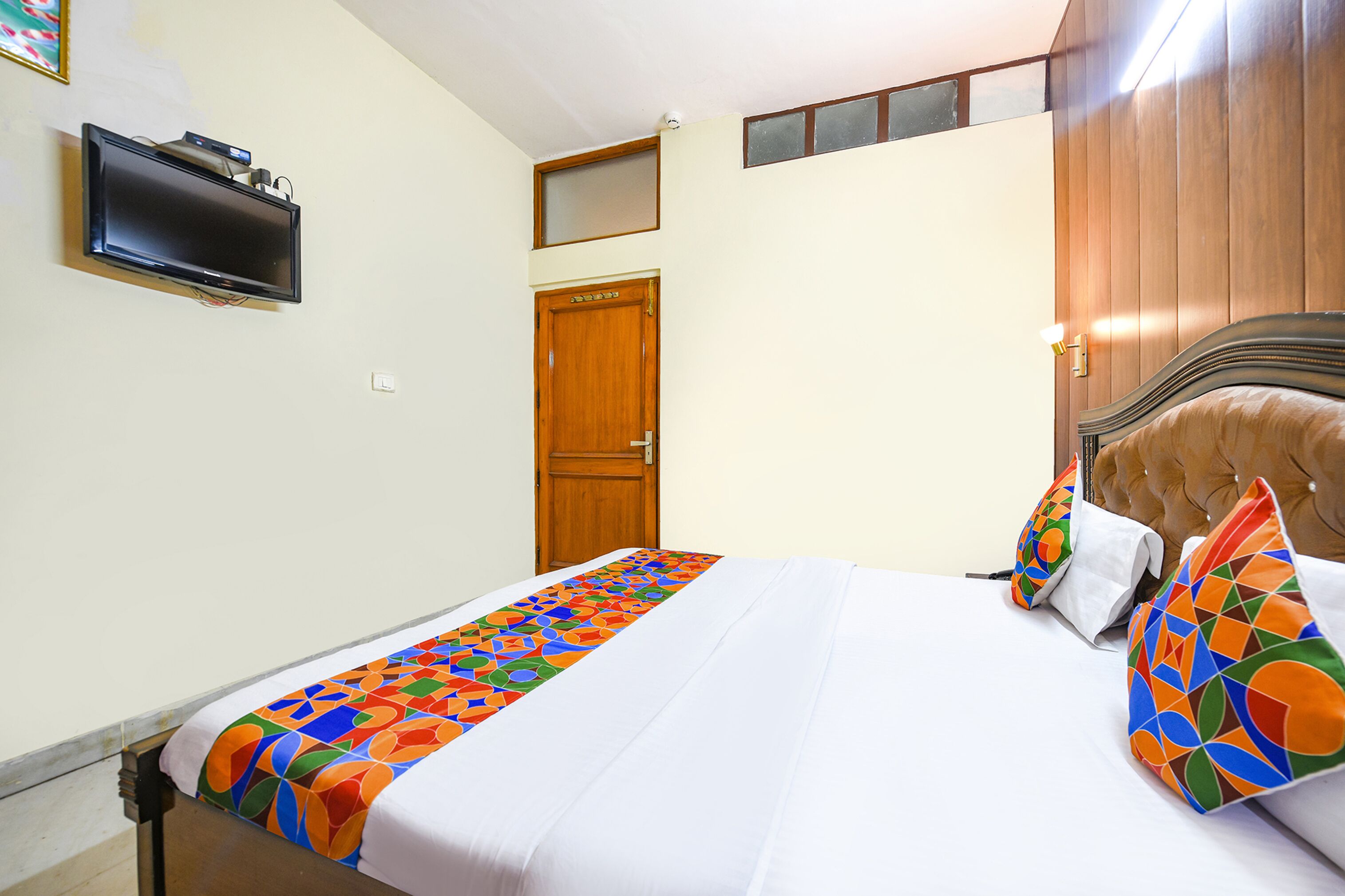 A hotel room with a bed, a TV mounted on the wall, a wooden door, and a window.