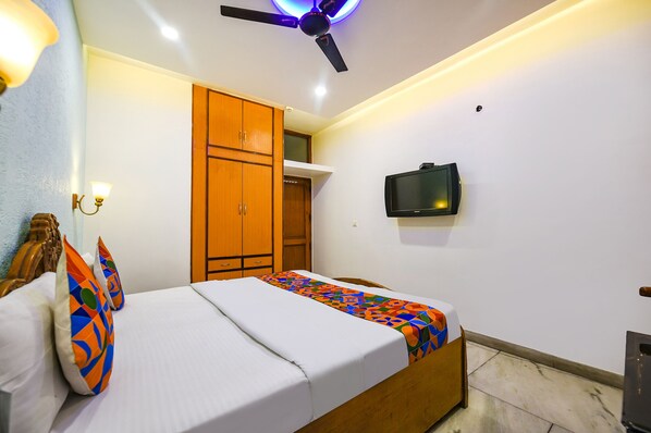 Une chambre d’hôtel comprenant un lit, une armoire en bois, un ventilateur de plafond, un téléviseur fixé au mur et une petite poubelle.