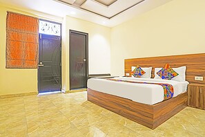 A bedroom with a wooden bed frame, a white mattress, and colorful pillows. There is a door with a curtain, a small cabinet, and a tiled floor. - FabHotel The Happiness (Kanpur)