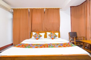 Quarto de hotel com cama de madeira, duas almofadas com padrões geométricos, colcha colorida, uma cadeira de madeira e uma mesa pequena.