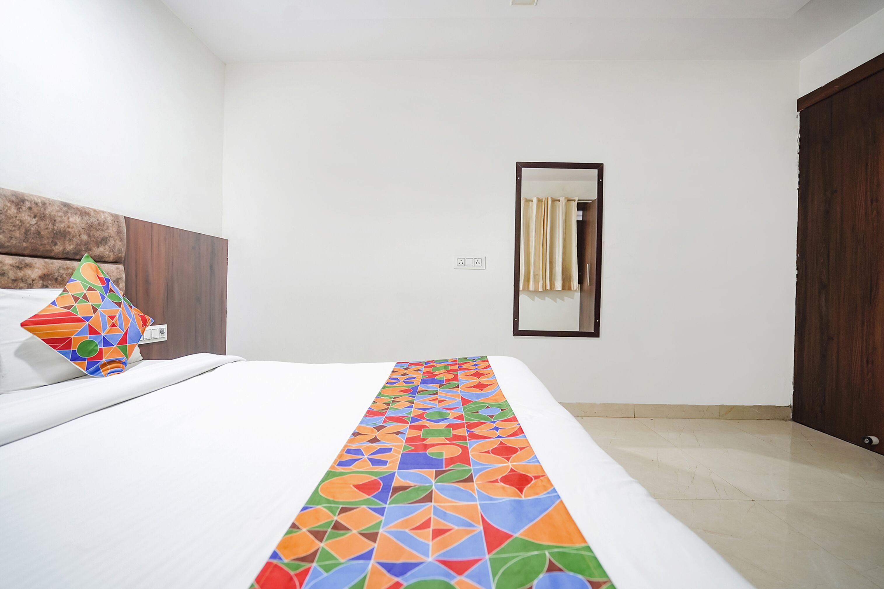 Ein Hotelzimmer mit einem Bett, einem bunten Läufer, einem hölzernen Kopfende, einem Spiegel und einer Tür.
