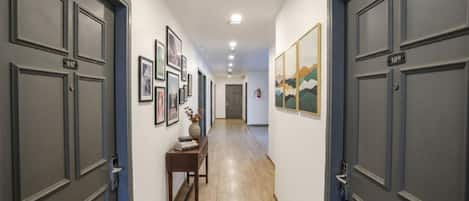 Un pasillo con puertas de color gris oscuro, con los números 107 y 109, que conducen a habitaciones. En las paredes hay cuadros enmarcados y una pequeña mesa con un jarrón y libros.