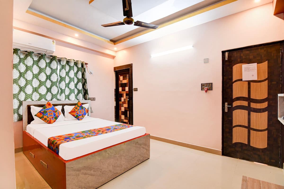 A bedroom with a bed, a ceiling fan, an air conditioning unit, a door with a decorative panel, and a small shelf.