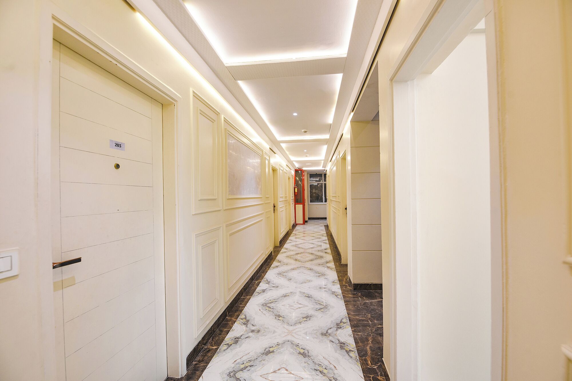 Un couloir d’hôtel avec des portes blanches, des panneaux indiquant le numéro des chambres et un sol en marbre à motifs.