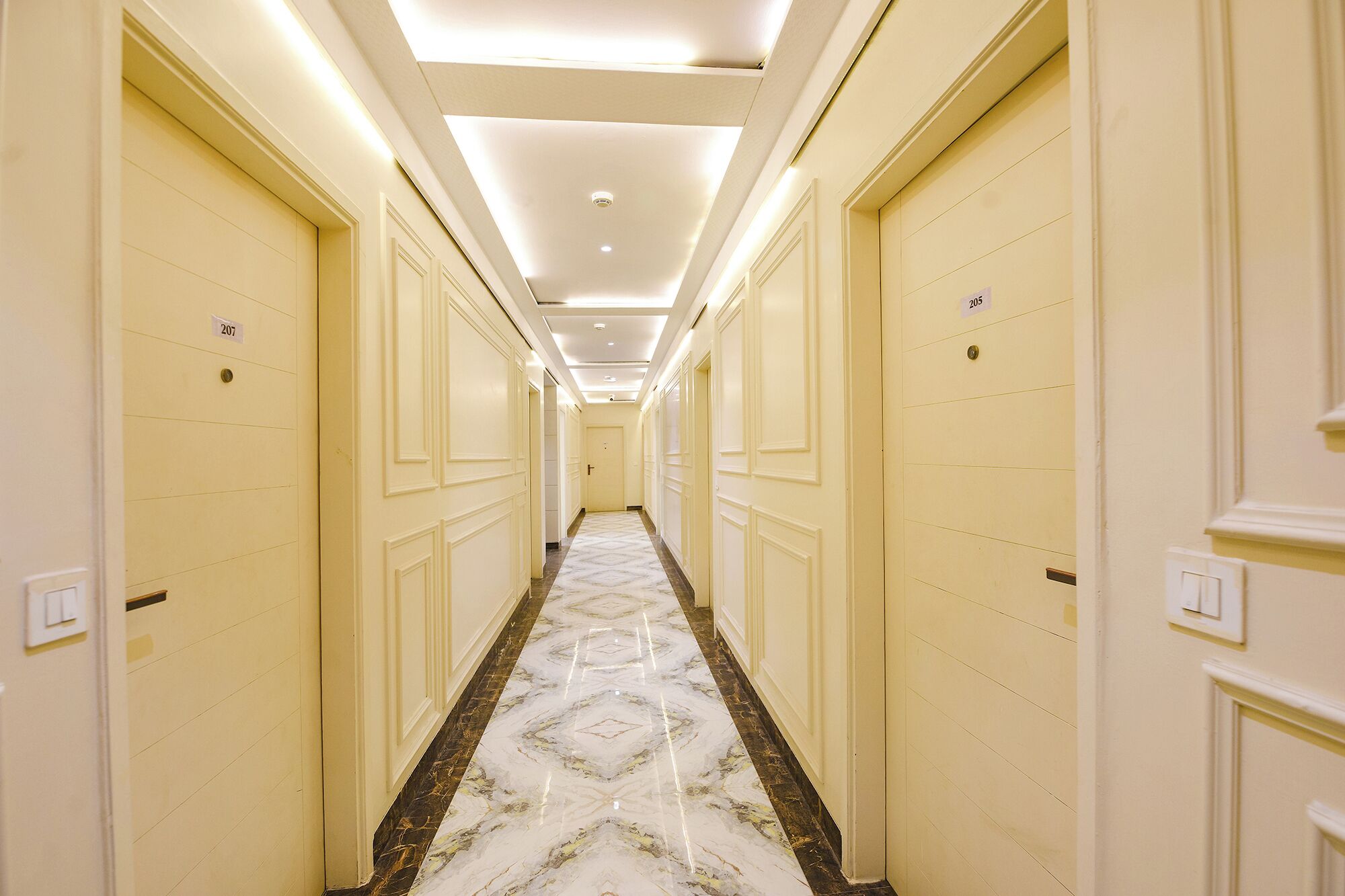 A long, narrow hallway with cream-colored walls and doors, marble flooring, and recessed lighting.