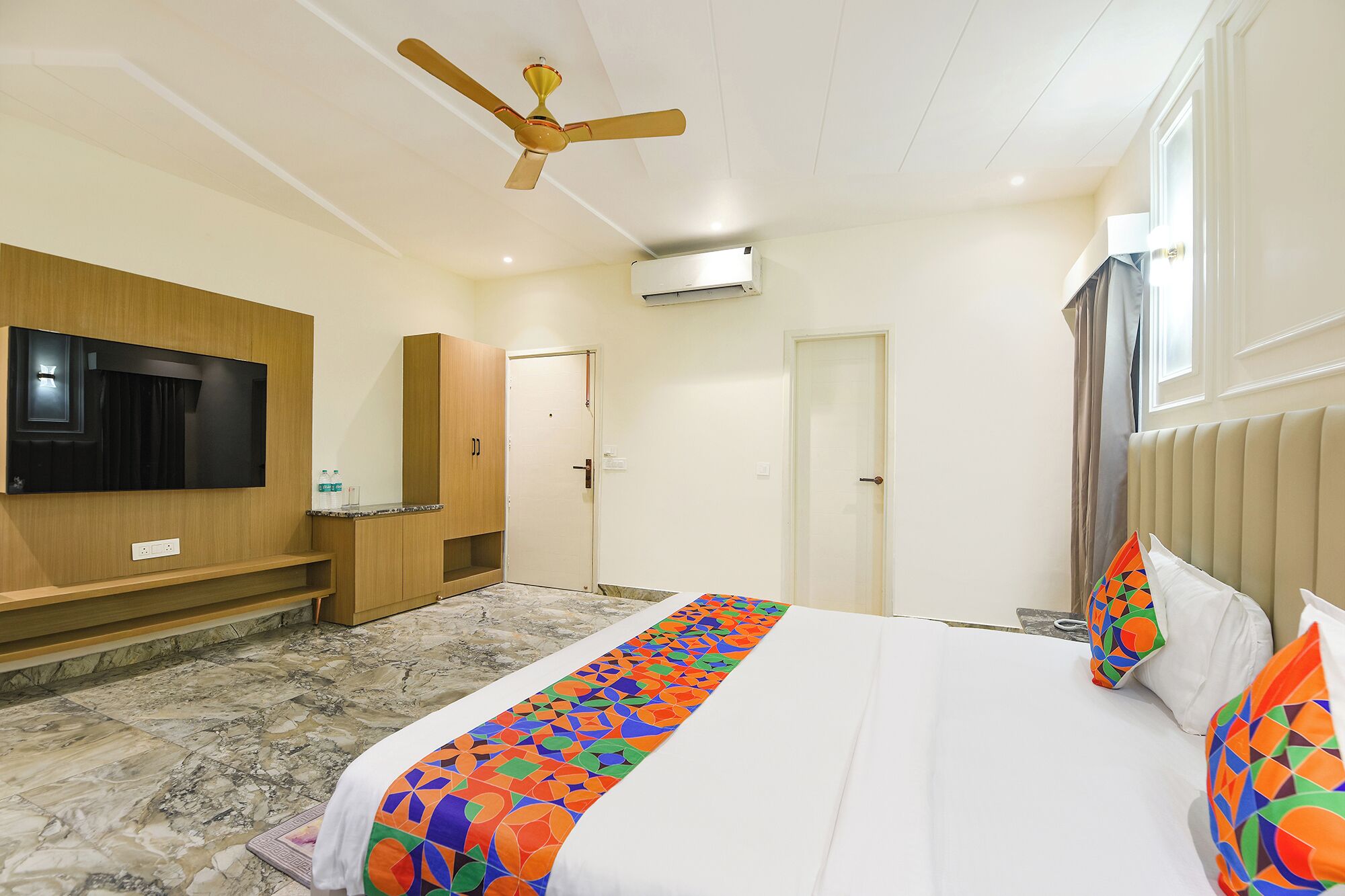 Una habitación de hotel con cama, televisor montado en la pared, ventilador de techo y ventana con cortinas.