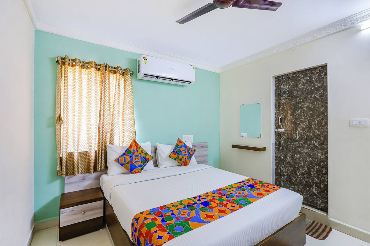 A bedroom with a bed, curtains, an air conditioning unit, a ceiling fan, a door with a marble-like pattern, and a small shelf.