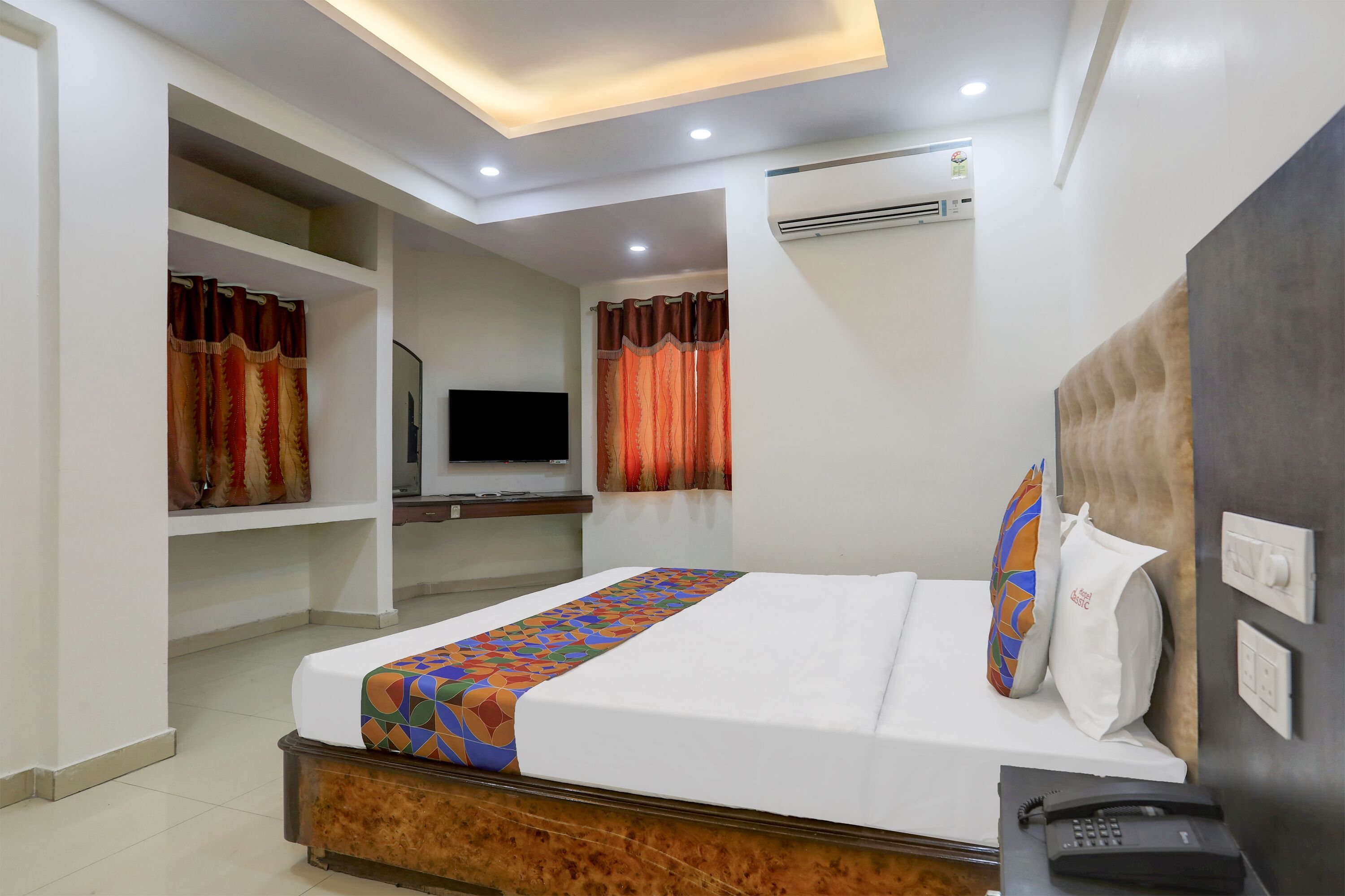 A hotel room with a bed, a TV, an air conditioner, and a telephone on a bedside table.