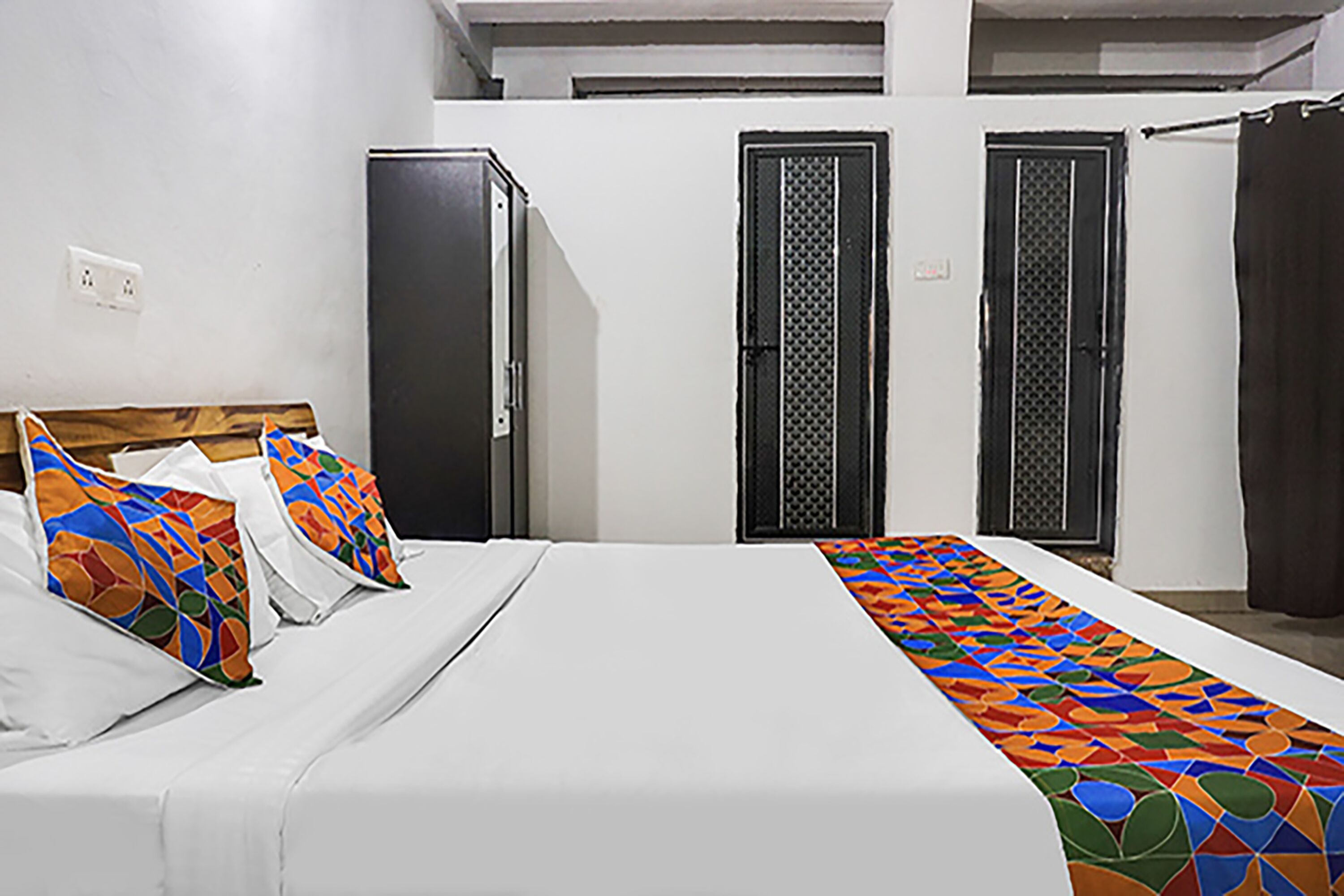 A bed with colorful pillows, a white bedspread, and a patterned runner. There are two doors and a curtain in the background.