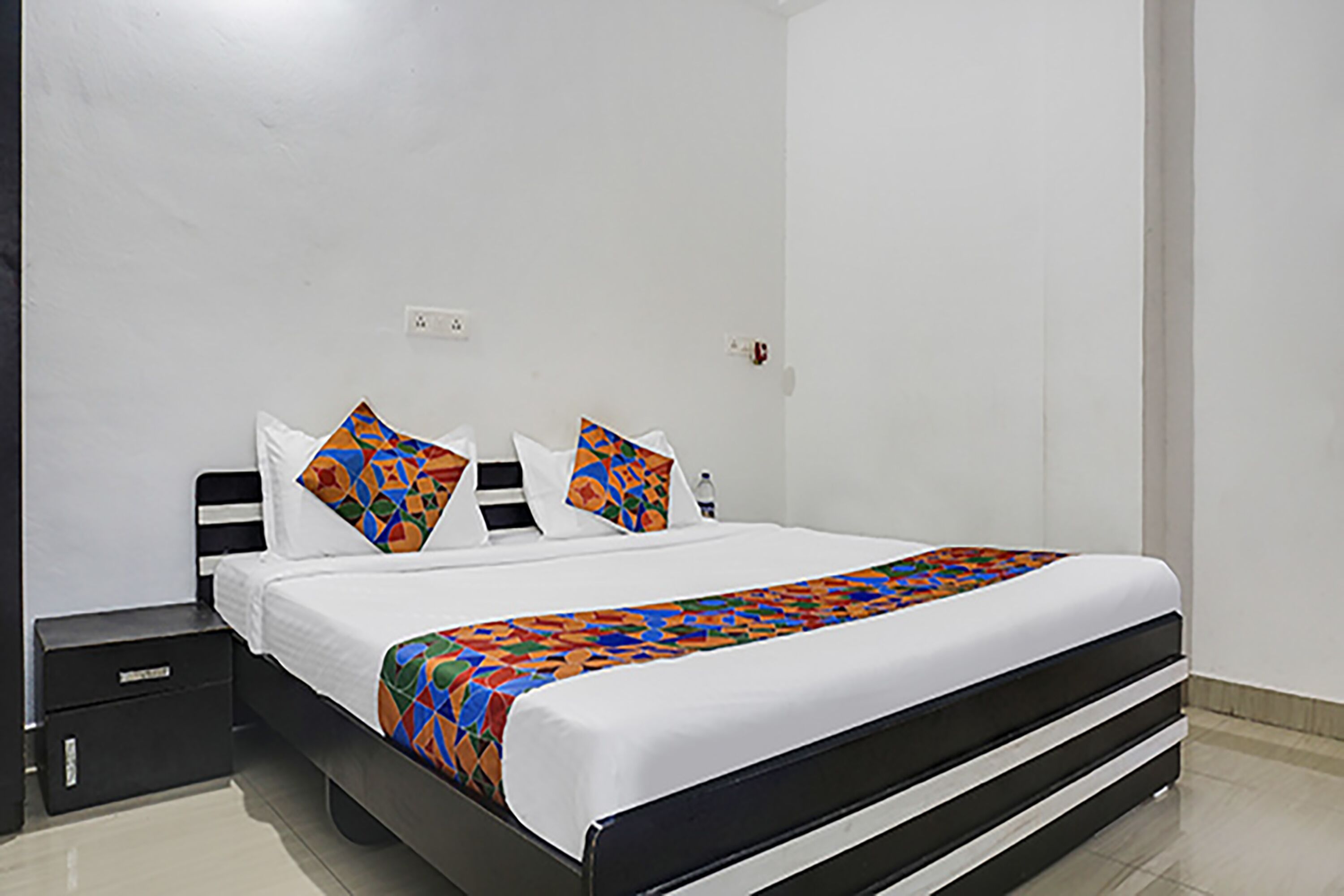 A neatly made bed with a colorful bedspread and two decorative pillows, a black nightstand with a drawer, and a white wall with a power outlet.
