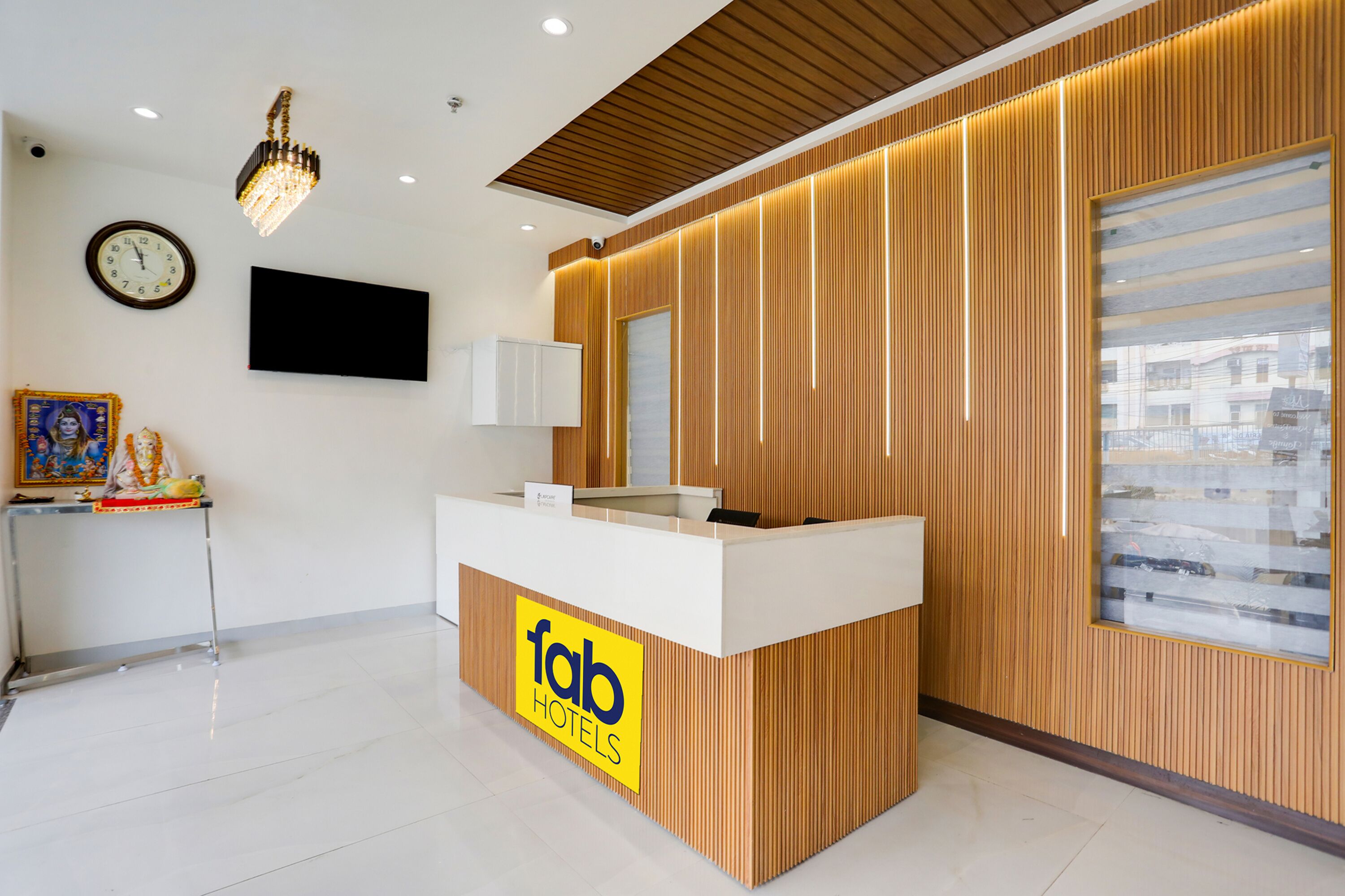A reception desk with a wooden finish and a 'fab hotels' logo. There is a wall-mounted clock and a flat-screen TV above the desk.