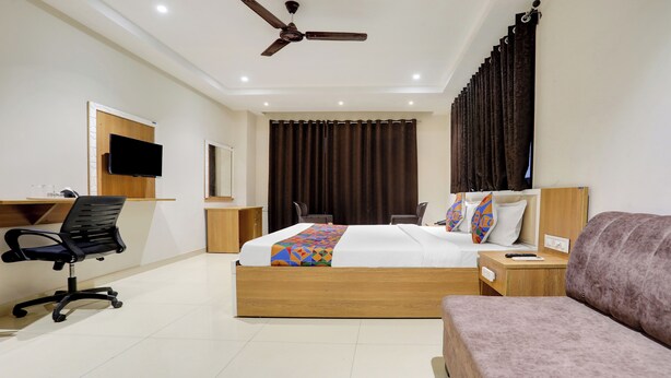 A hotel room with a bed, a desk with a chair, a sofa, a ceiling fan, and a TV on the wall. - FabHotel Mansarovar Inn (Raipur)