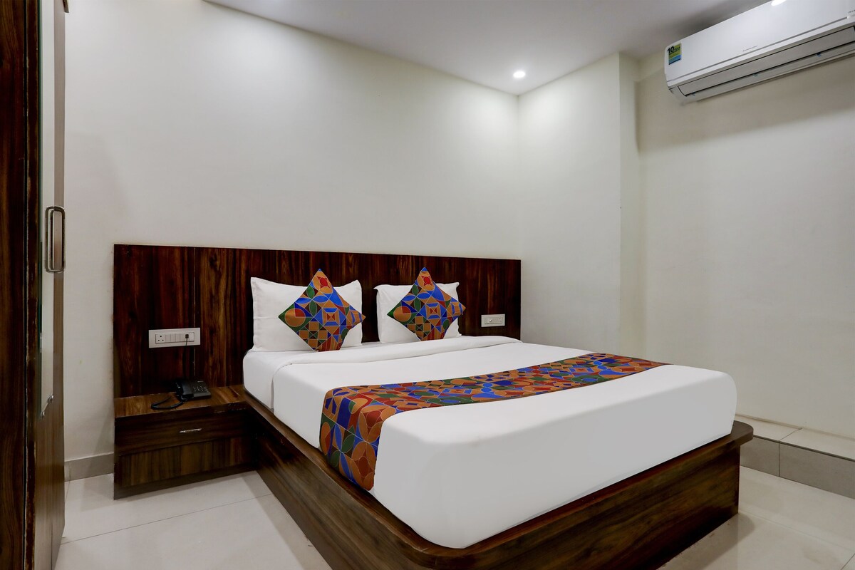 Habitación de hotel con cabecera de madera, cama blanca con almohadas coloridas, mesita de noche con teléfono y unidad de aire acondicionado en la pared.