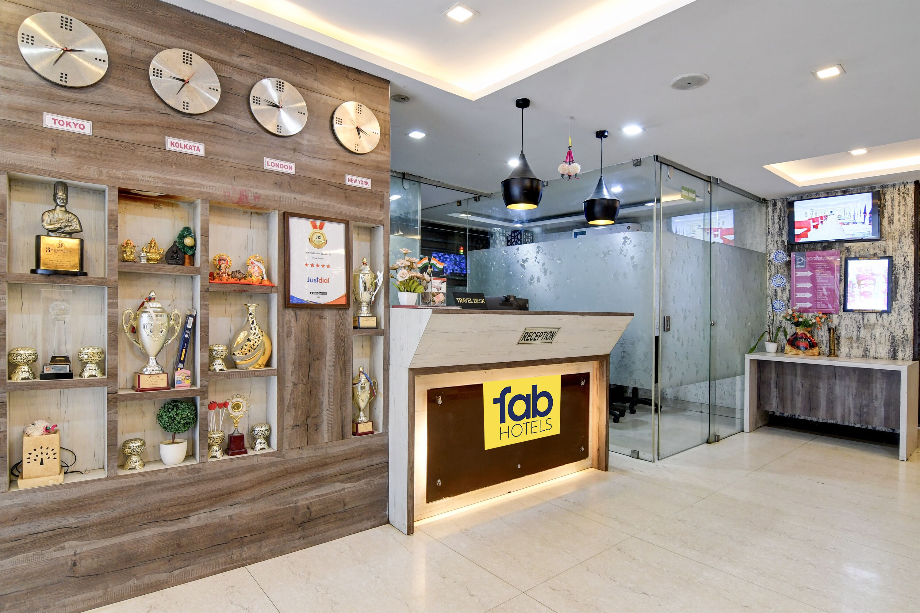 A hotel reception area with a wooden display of trophies and clocks, a reception desk, and a glass wall.