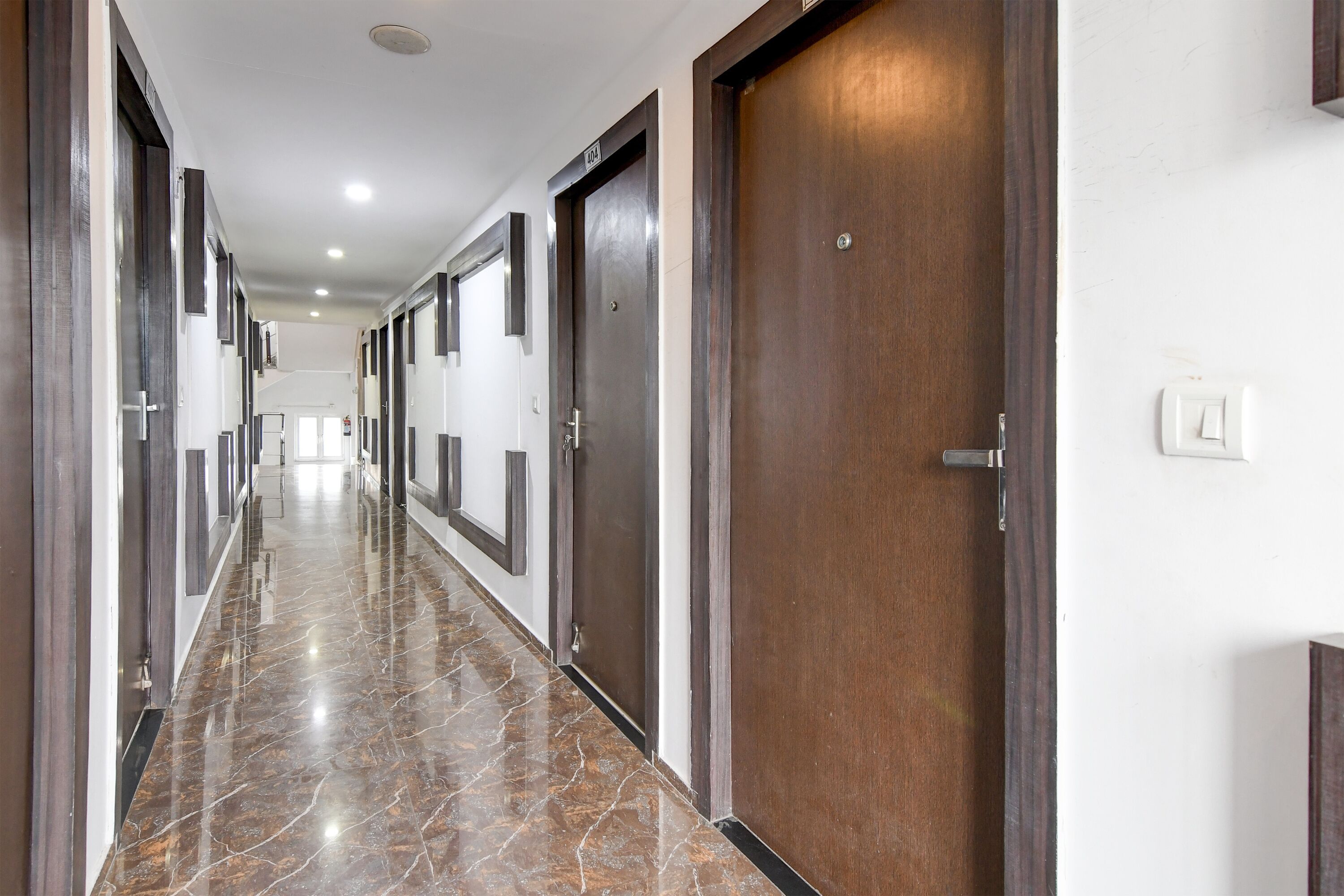 A long, well-lit hallway with marble flooring and wooden doors, leading to a bright room at the end.