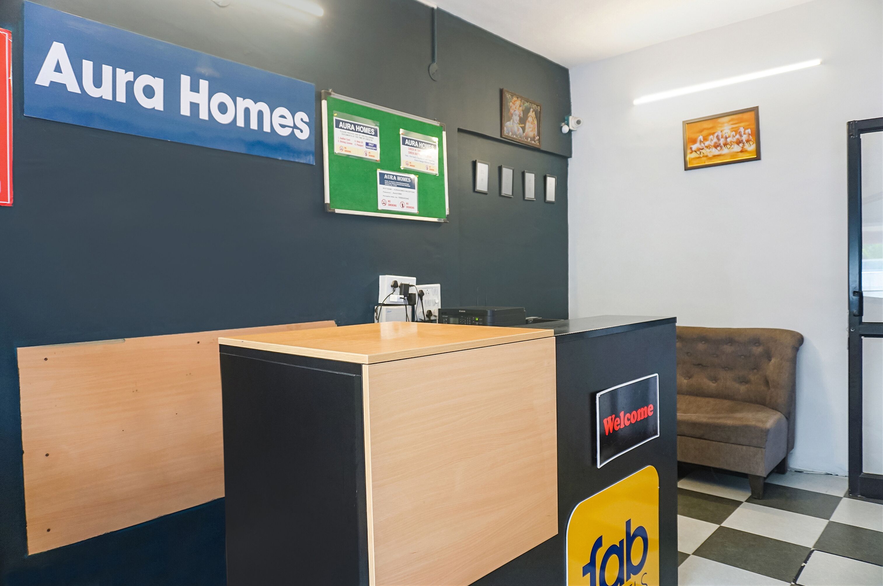 Un mostrador de recepción con un cartel de 'Welcome', un letrero de 'Aura Homes' y una pizarra verde con avisos.
