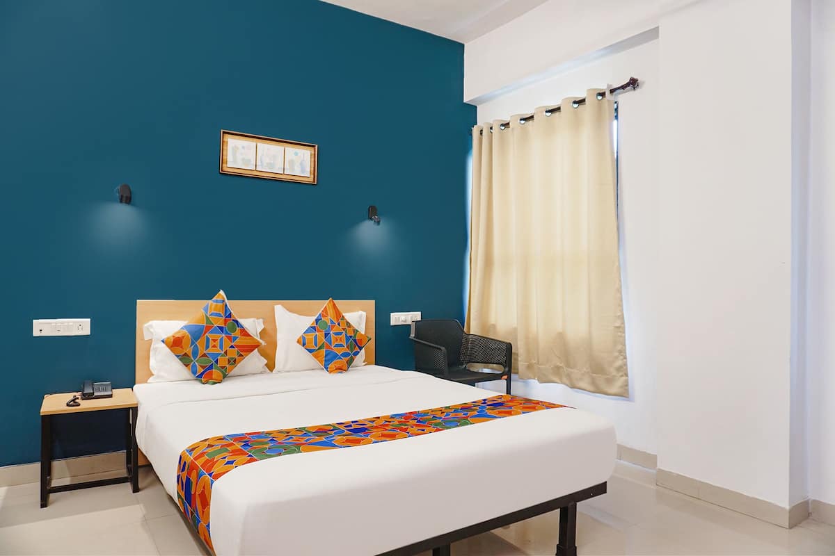 Habitación de hotel con una cama grande, una mesita de noche, una silla, una ventana con cortinas y una lámpara fijada a la pared.