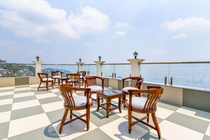 Une terrasse sur le toit, dotée d’un sol à carreaux, de chaises en bois avec des coussins et d’une rambarde en verre offrant une vue sur le paysage urbain.