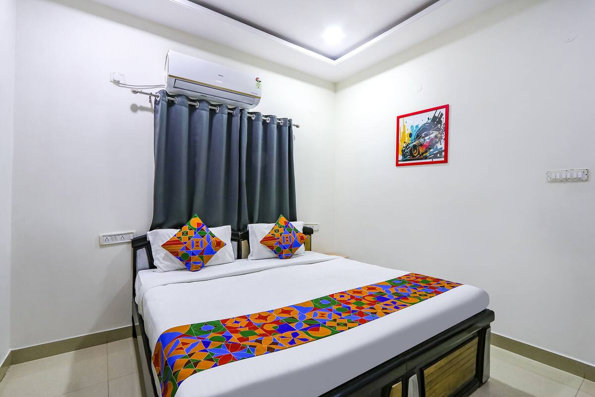 Una habitació d'hotel amb llit, aire condicionat, cortines, un estovat de llit colorit i un quadre a la paret.