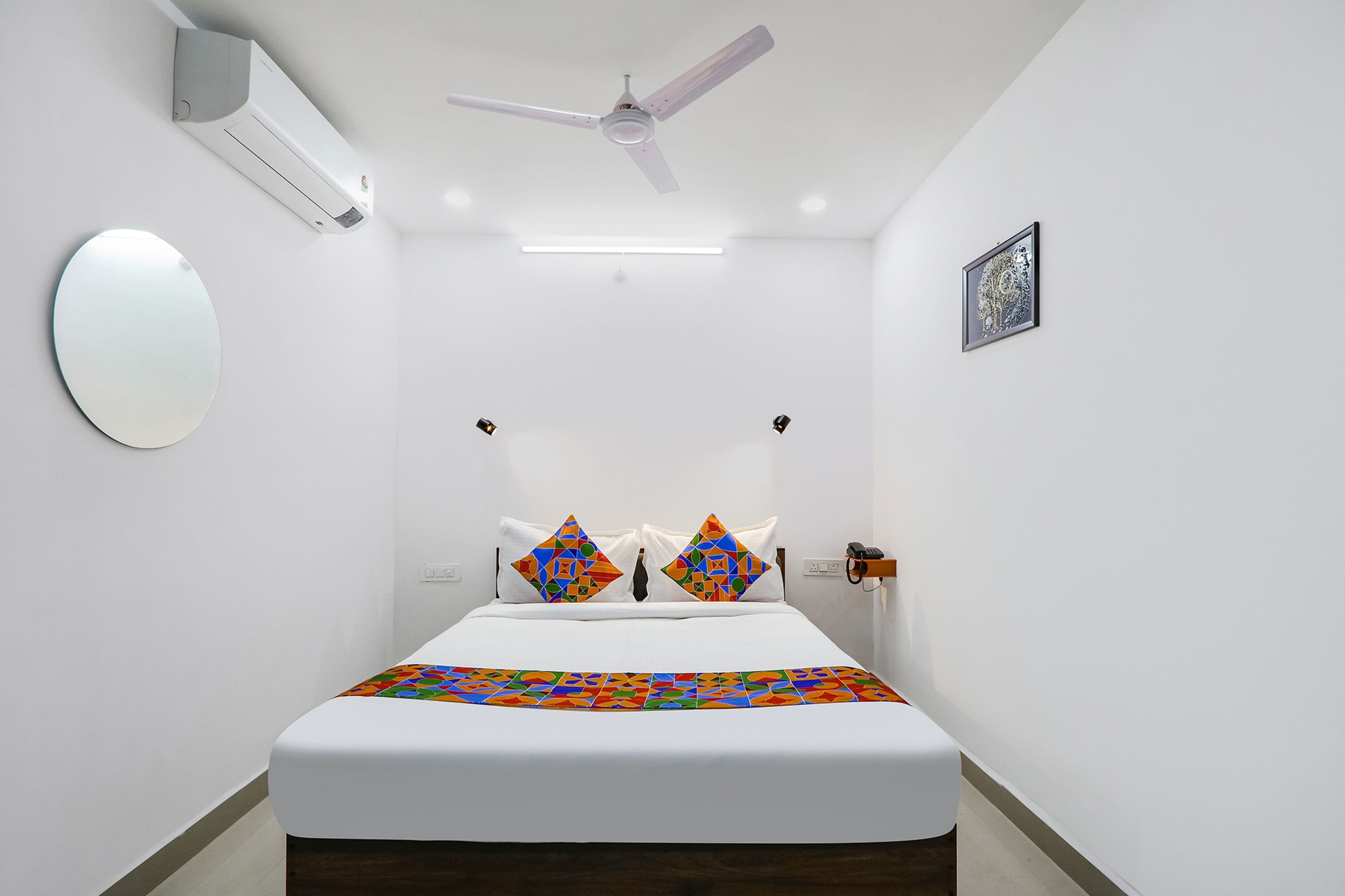 A modern hotel room with a bed, a ceiling fan, an air conditioning unit, a bedside table with a lamp, and a framed artwork on the wall.