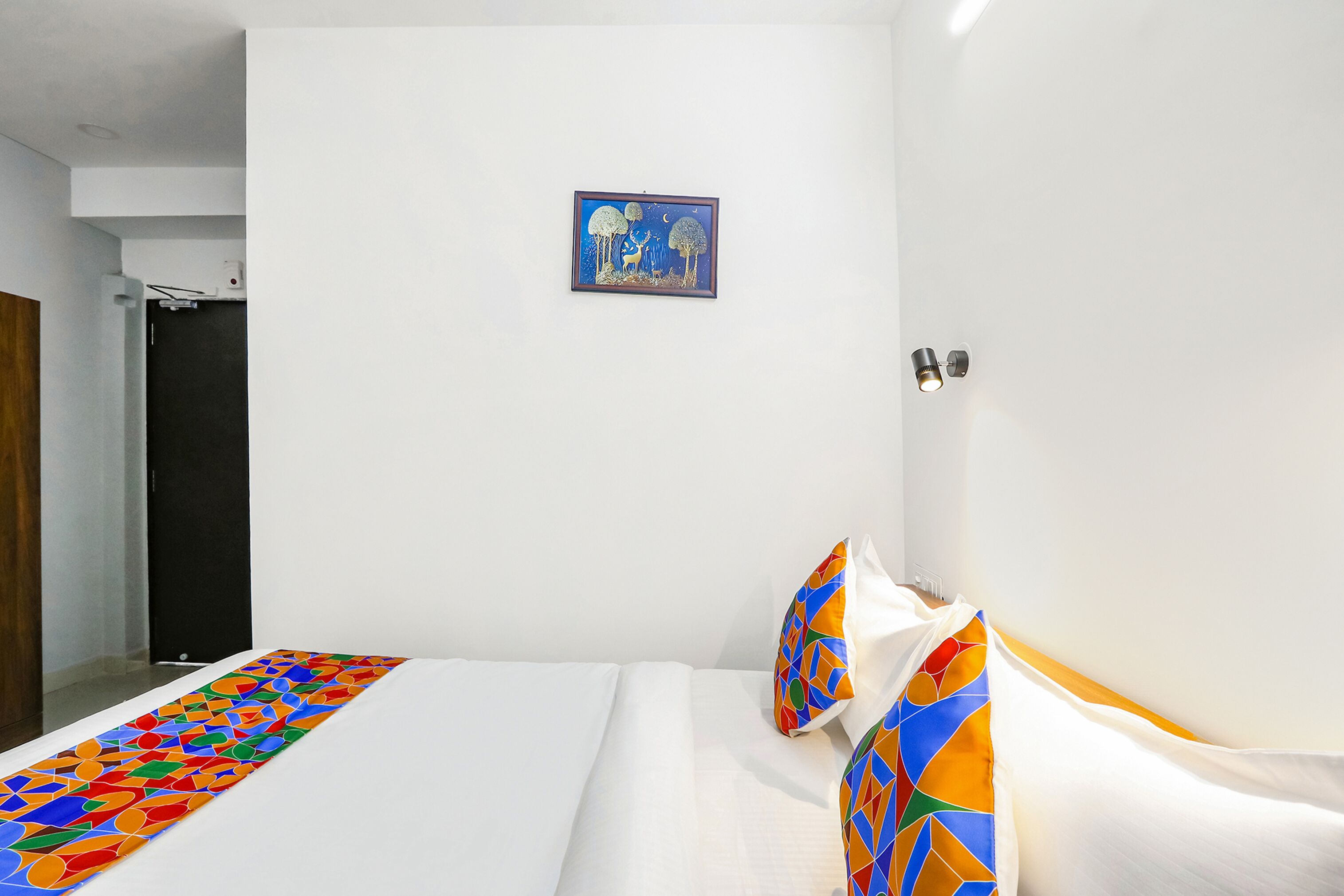 Ein Hotelzimmer mit einem Bett, einer farbenfrohen, geometrisch gemusterten Tagesdecke, einem gerahmten Kunstwerk an der Wand und einer Tür links.