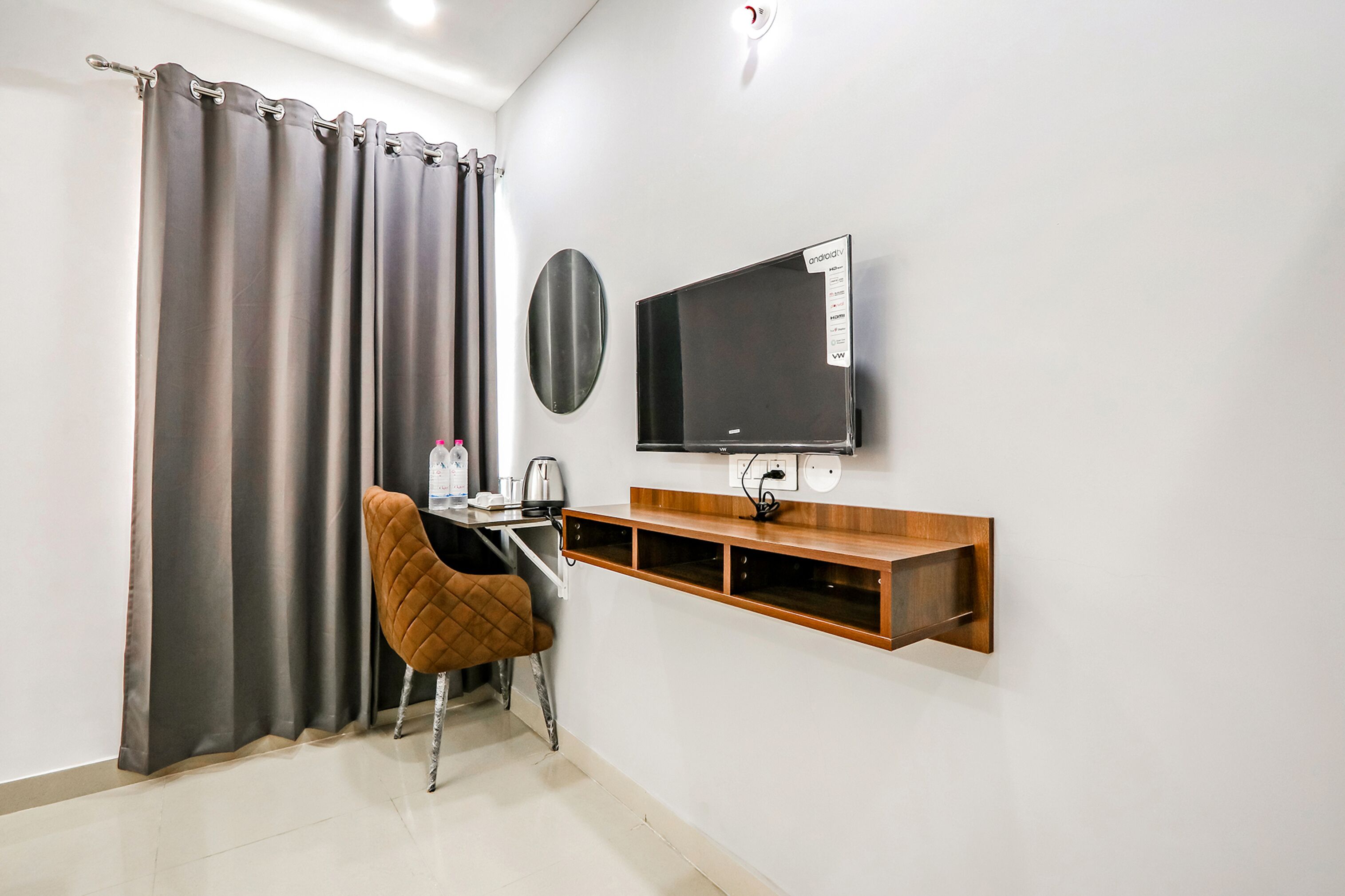 A modern room with a flat-screen TV mounted on the wall, a wooden shelf below it, a chair, and a curtain.