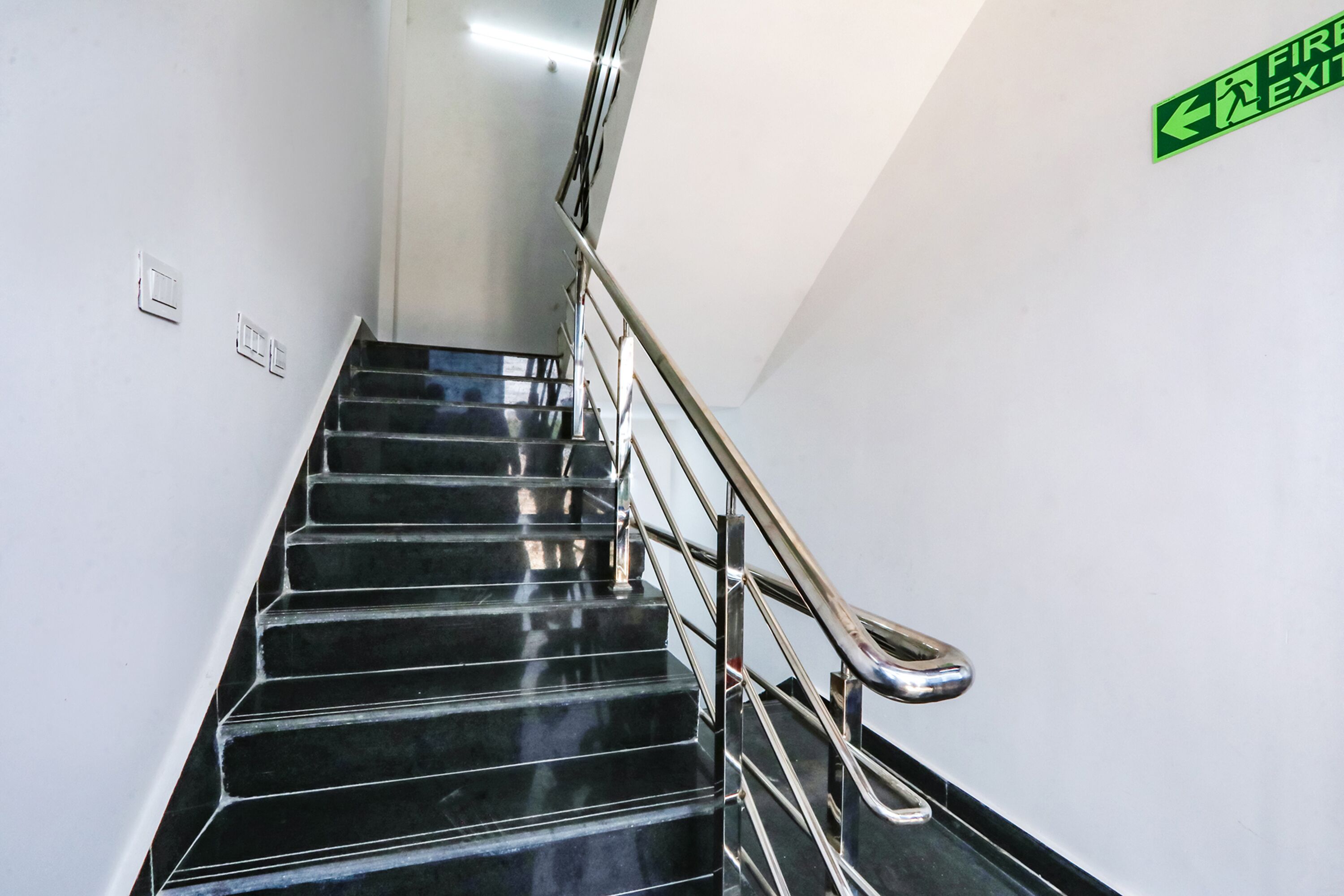 Una escalera con peldaños negros y barandilla metálica, un letrero de salida de incendios en la pared, y un interruptor de luz en la pared.