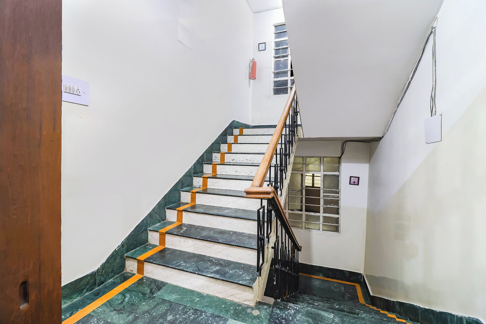 A staircase with marble steps and wooden handrails, leading to a landing with a window and a fire extinguisher.