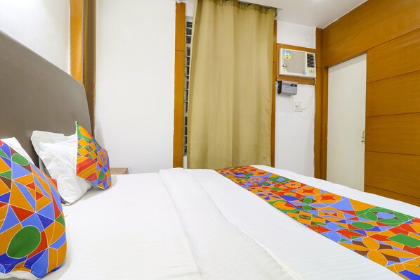 A hotel room with a bed, colorful pillows, a window with curtains, an air conditioning unit, and a wooden door. - FabHotel Nand (Patna)