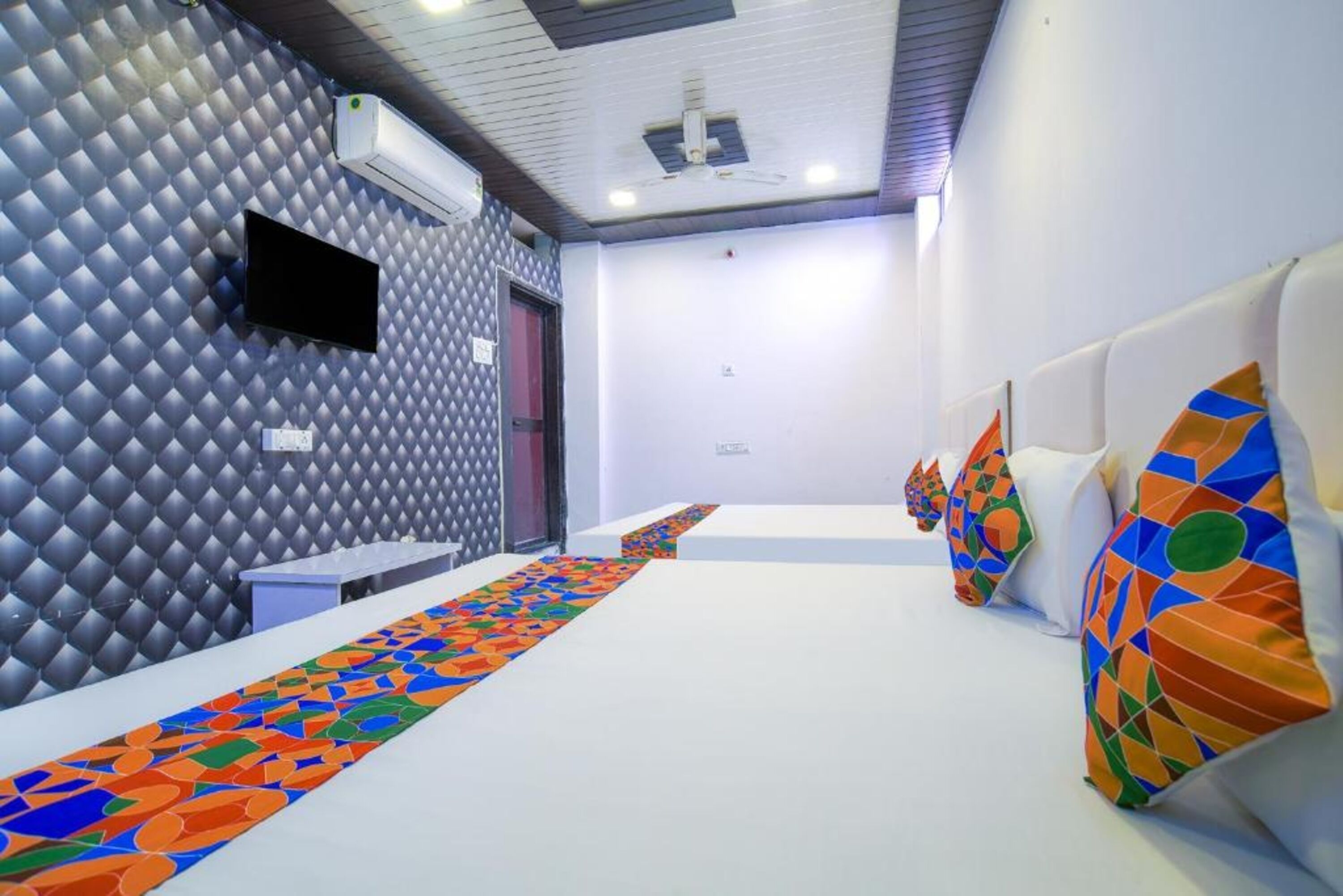 Habitación de hotel con pared estampada, televisor de pantalla plana, alfombra larga con diseño y una fila de camas con almohadas de colores.