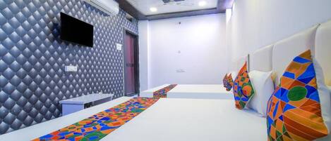Habitación de hotel con pared estampada, televisor de pantalla plana, alfombra larga con diseño y una fila de camas con almohadas de colores.
