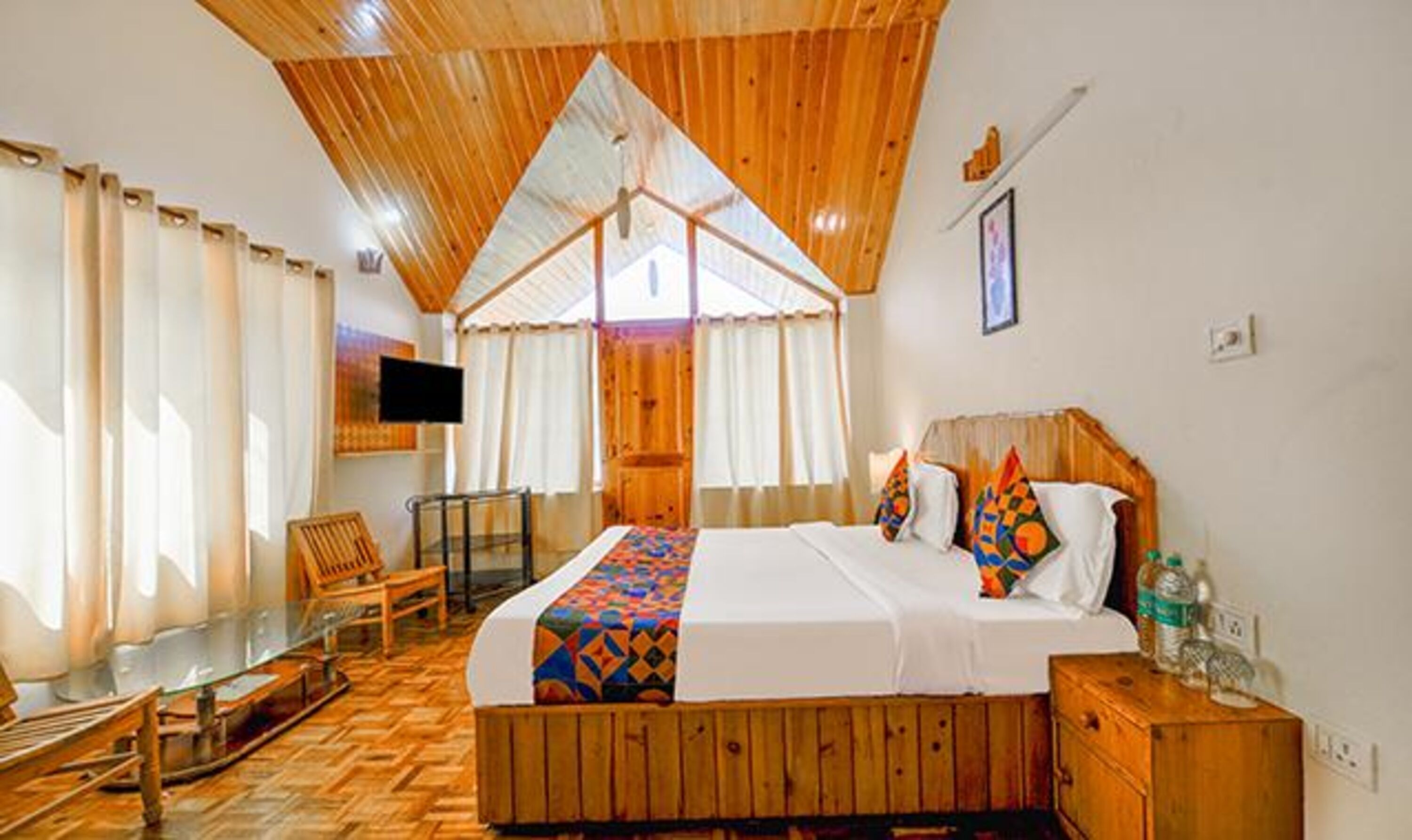 A hotel room with a wooden ceiling, a bed with colorful bedding, a wooden nightstand, a chair, and a TV mounted on the wall.