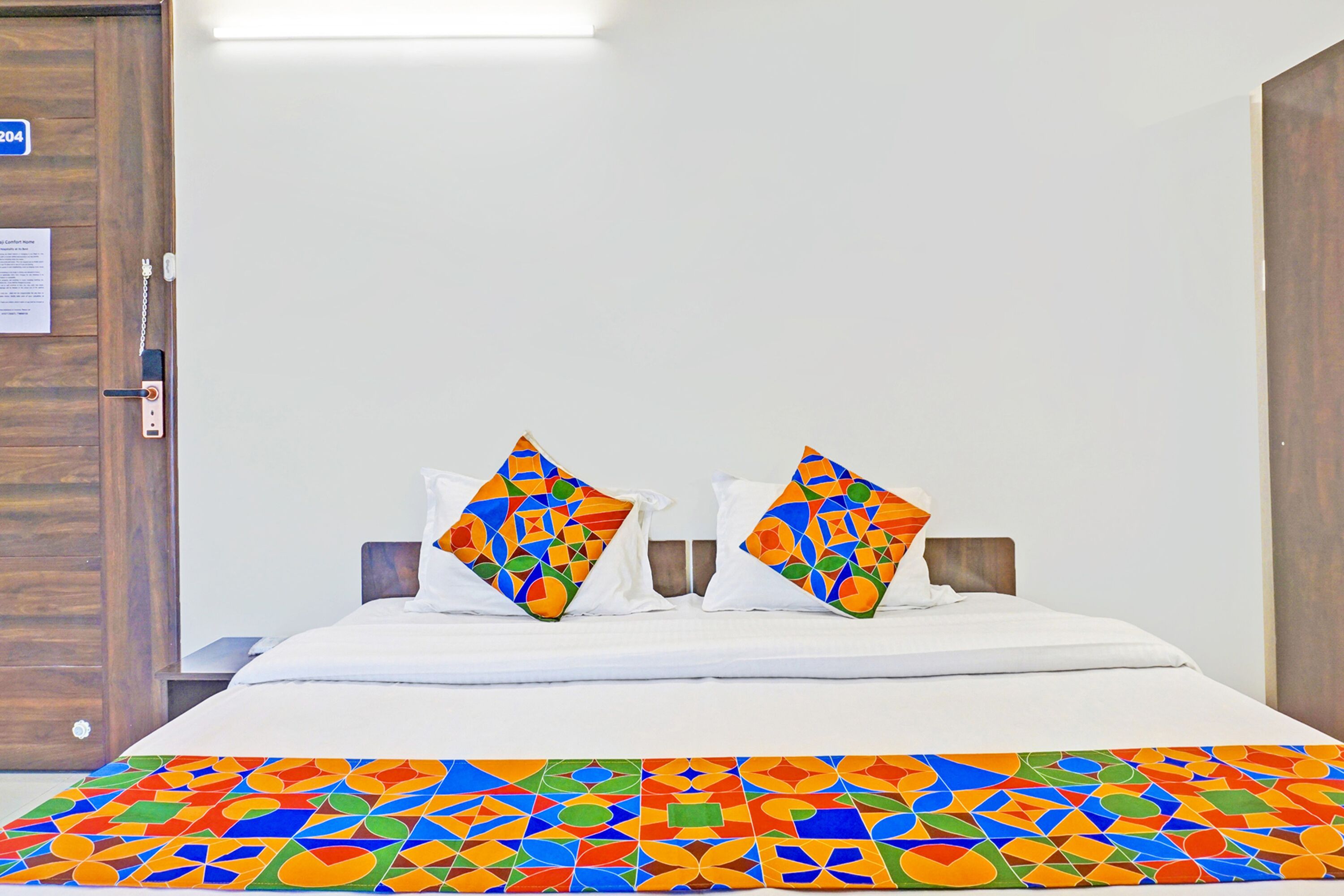 Una habitación de hotel con una cama, dos almohadas de estampado geométrico colorido y una colcha blanca, junto con un plaid de diseño geométrico y colorido.