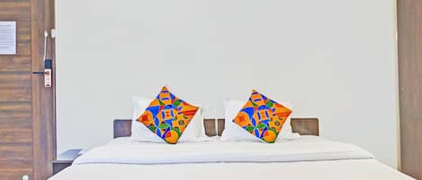 Una habitación de hotel con una cama, dos almohadas de estampado geométrico colorido y una colcha blanca, junto con un plaid de diseño geométrico y colorido.