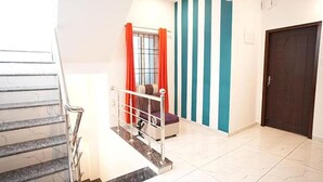 Una escalera con barandilla metálica, una cama con cubrecamas estampado, una ventana con cortinas naranjas y una puerta con rayas verticales azul turquesa y blanca.