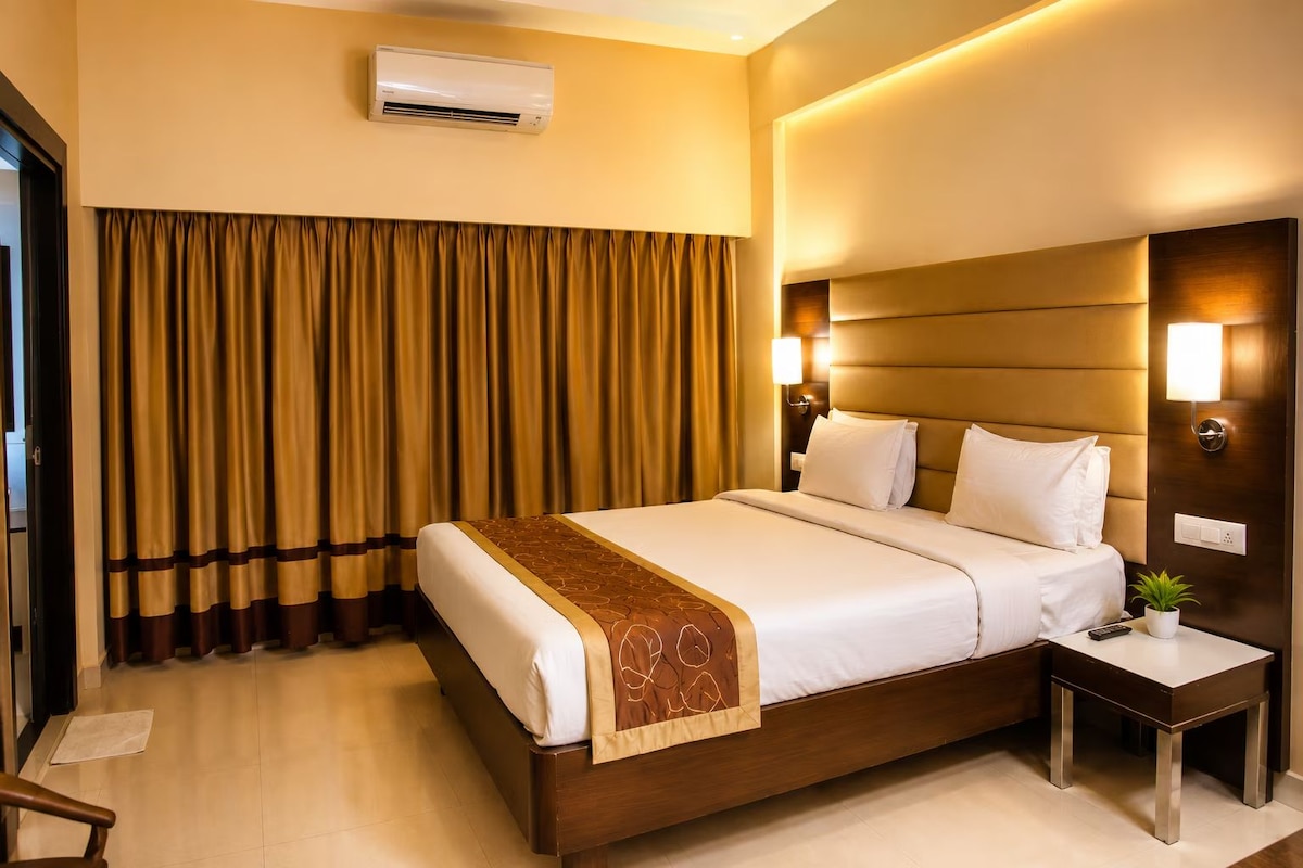 A hotel room with a large bed, a bedside table, an air conditioning unit, and a window with curtains.