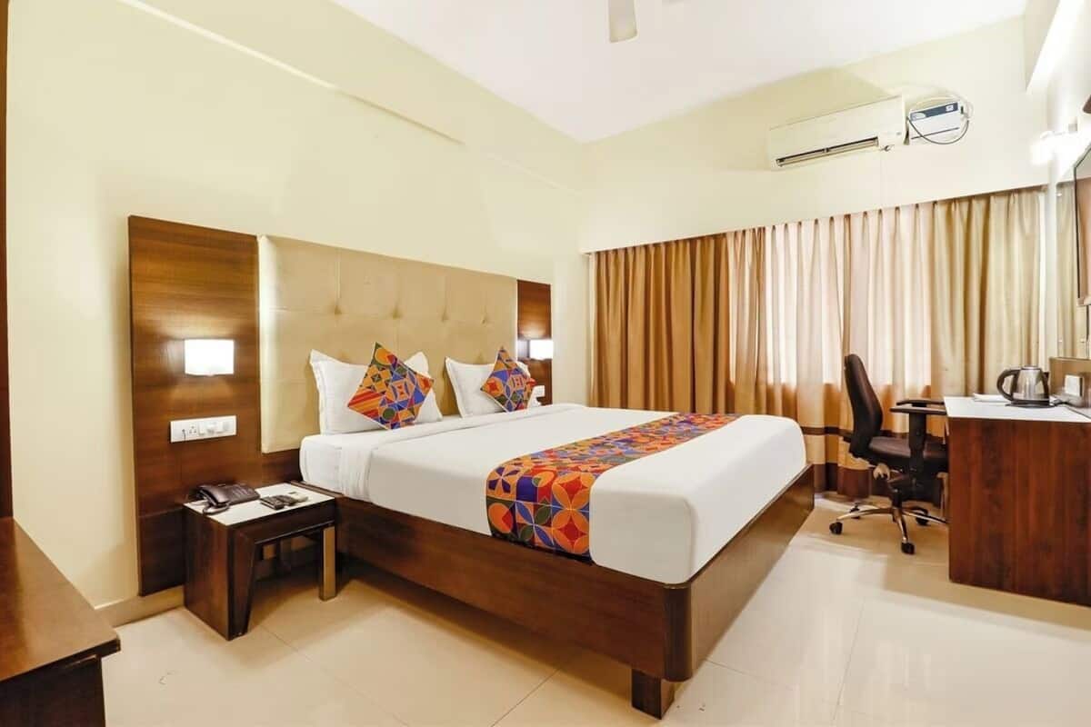 A hotel room with a large bed, a desk with a chair, an air conditioning unit, and a window with curtains.