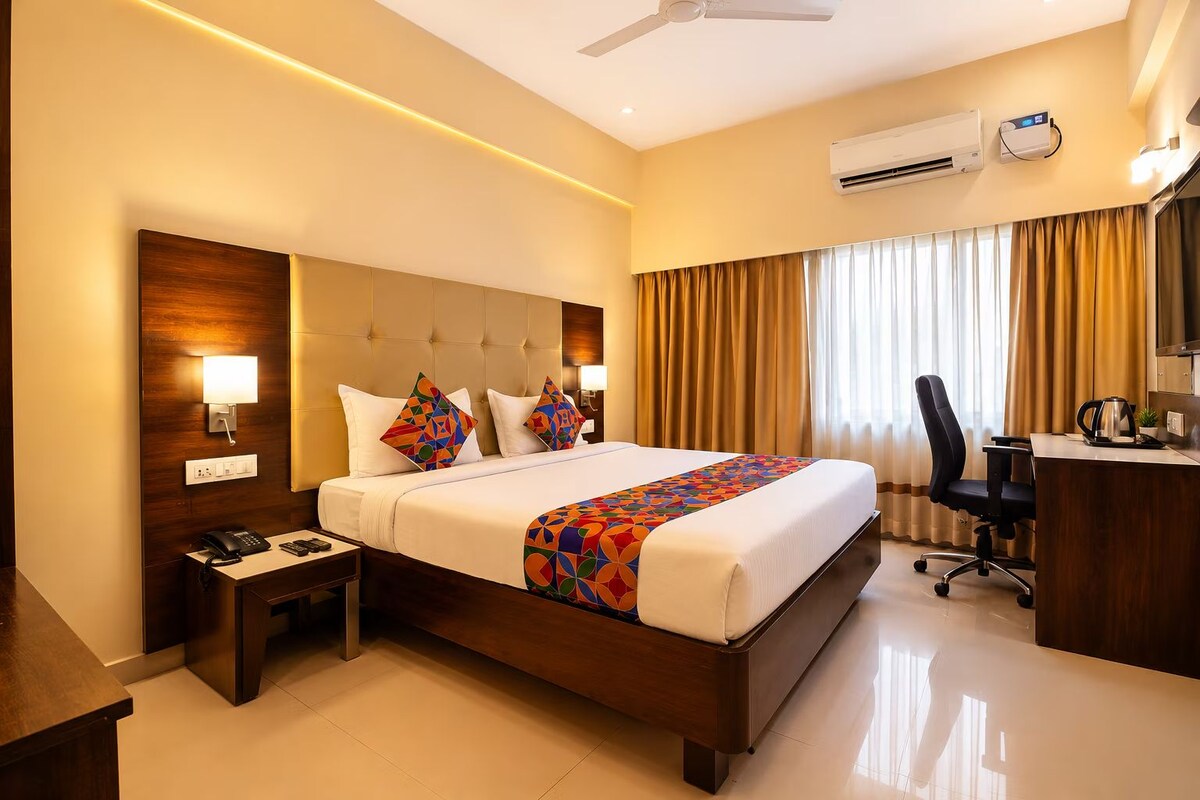 A hotel room with a bed, bedside table, lamp, air conditioning unit, and a desk with a chair.