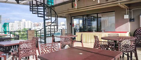 An outdoor dining area with multiple tables and chairs, a spiral staircase, and a view of the cityscape in the background.