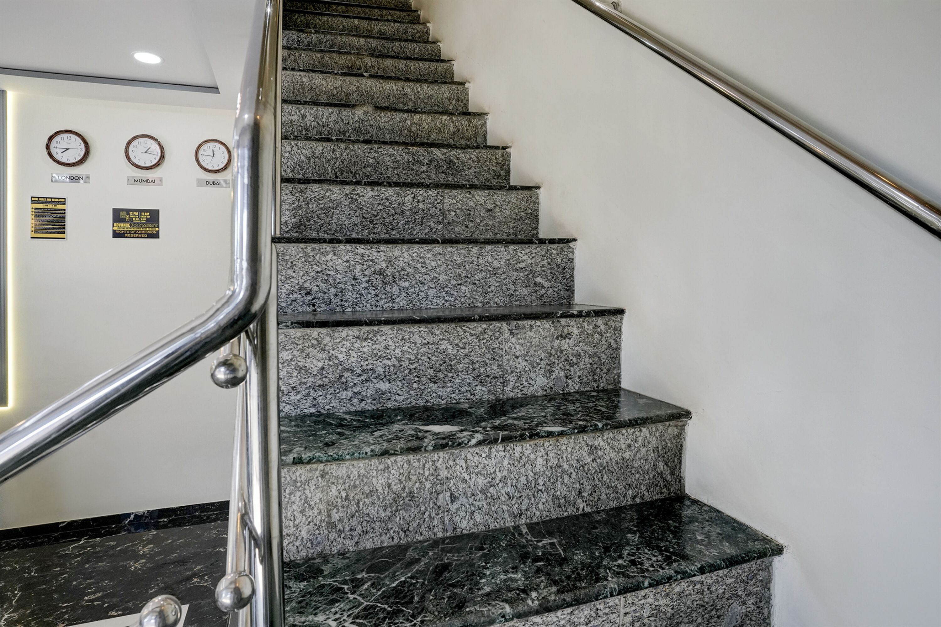 A staircase with granite steps and a metal handrail, leading to a landing with clocks and signs indicating time zones.
