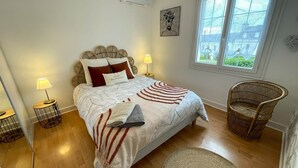 Quarto com cama, duas luminárias de cabeceira, uma cadeira de vime, uma janela e uma obra de arte na parede.