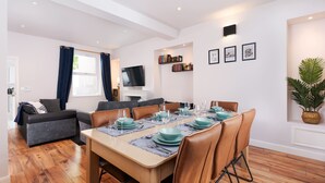 Ein modernes Wohnzimmer mit Essbereich, Holzboden, einem Flachbildfernseher, einem Bücherregal und einer Topfpflanze.