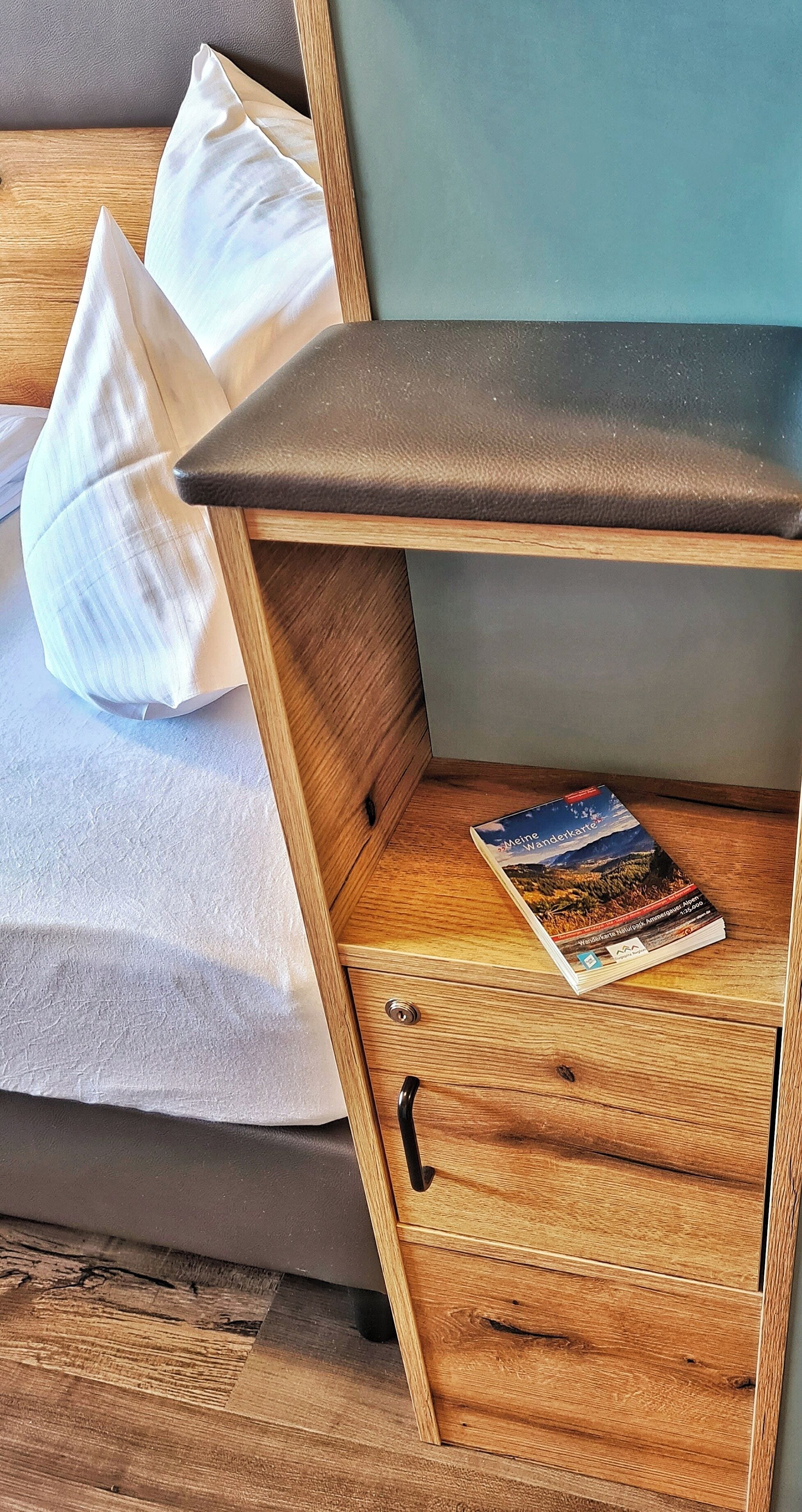 A bed with pillows, a wooden nightstand with a book, and a drawer with a pen.
